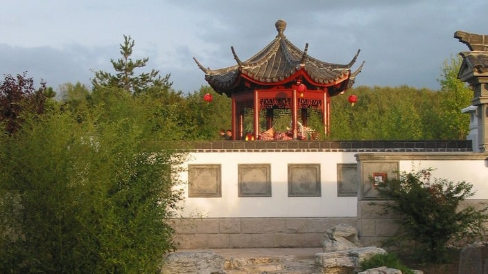 Blick in den Chinesischen Garten: Im Vordergrund unregelmäßige Steinplatten, im Hintergrund ein rot lackierter Pavillon.