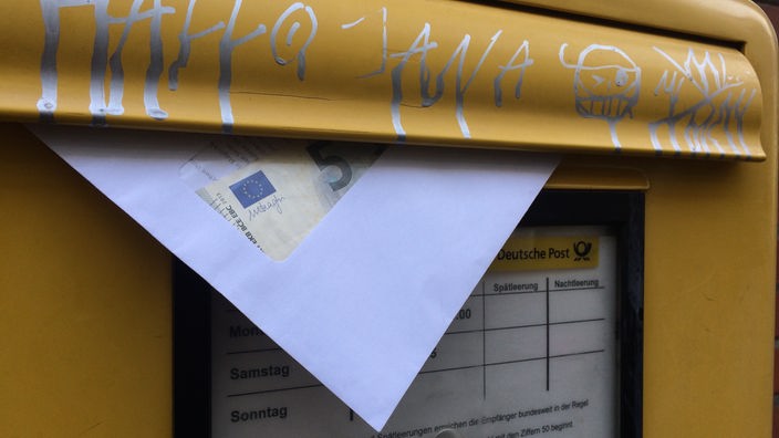 Briefumschlag hängt aus einem Briefkasten heraus, der mit Graffiti beschmiert ist; im Sichtfenster des Kuverts ist ein 5-Euro-Schein zu erkennen