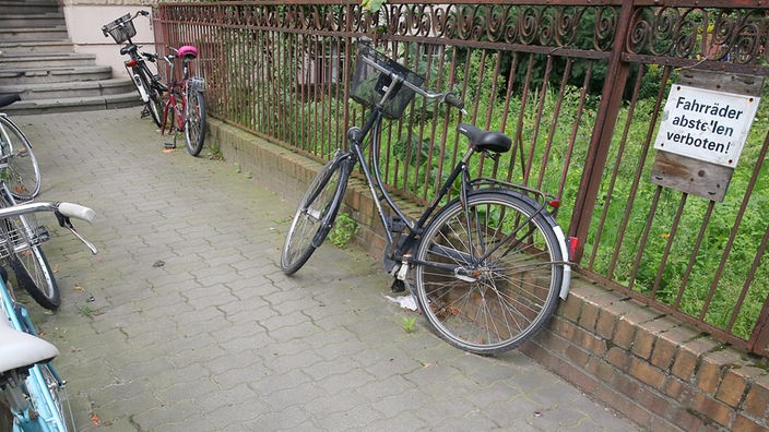 Fahrräder lehnen an einem Zaun, an dem ein Schild mit der Aufschrift "Fahrräder abstellen verboten" hängt