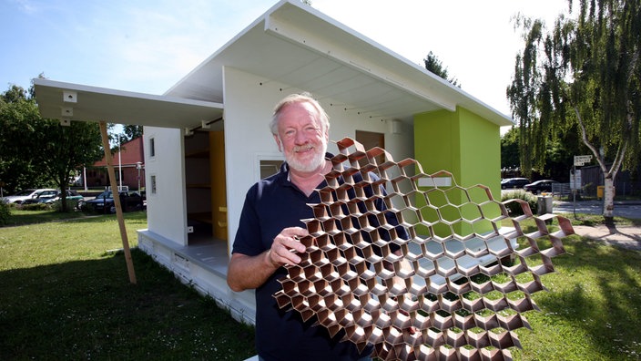 Ein Mann hält eine Pappe in Wabenform vor einem Haus aus Papier hoch