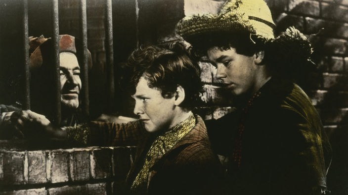 Filmszene: Tom Sawyer und Huckleberry Finn besuchen einen Gauner, der durch ein vergittertes Fenster zu den Jungen herausspäht.
