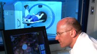 Ein Arzt analysiert ein PET-CT-Bild; im Hintergrund ist die Röhre für diese Positronen-Emissions-Tomographie zu erkennen.