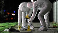 Beamte der Spurensicherung stellen auf einem Gehweg Nummerntafeln auf. Sie tragen Schutzkleidung.