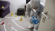 Ein Beamter in Schutzkleidung sucht nach Fingerabdrücken. Auf dem Boden sind Blutspuren und verschiedene Beweise.