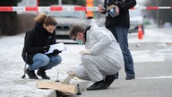 Eine Polizistin hockt neben einem Beamten der Spurensicherung. In der Hand hält sie ein Klemmbrett.