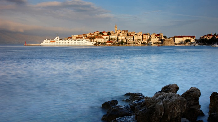 Blick übers Meer auf die historische Altstadt von Korčula, an der eine weiße Fähre vorbeifährt. | Bildquelle: Interfoto Blick übers Meer auf die historische Altstadt von Korčula, an der eine weiße Fähre vorbeifährt.