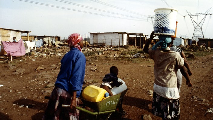 Wellblechhütten eines Slums, davor schwarze Frauen mit Wasserkanistern