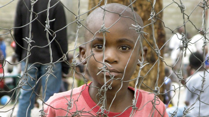 Ausschnitt eines Stacheldrahtzaunes, dahinter ein Kind in einem Flüchtlingslager