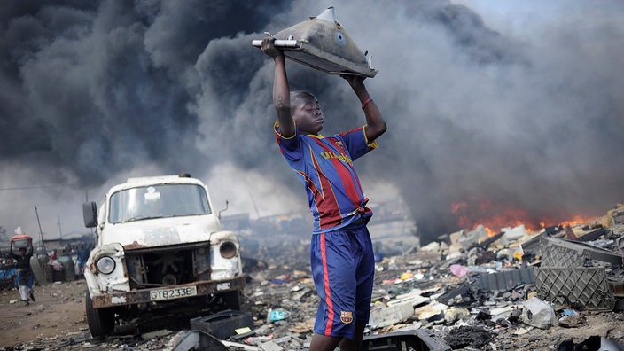 Ein Junge hebt auf der Giftmüllhalde Agbogbloshie in Ghana einen alten Monitor nach oben, hinter ihm toxischer Rauch.