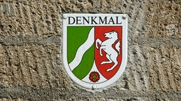 In einer alten Mauer ist eine Plakette eingelassen, auf der der Schriftzug Denkmal sowie das Wappen von NRW zu sehen sind. 