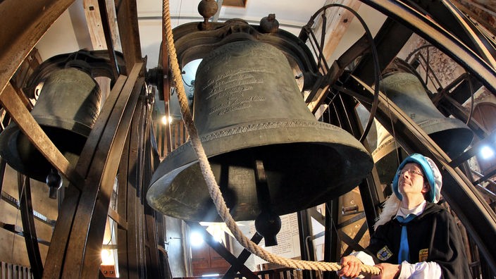 Im Vordergrund des Glockenturms ist eine große Bronzeglocke, im Hintergrund sind zwei weitere Glocken zu sehen, ein Mann zieht an einem Seil.
