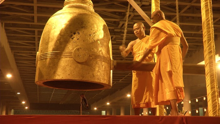 Zwei buddhistische Mönche bringen mit einem großen Klöppel eine goldene Glocke zum Schwingen