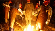 Glockengiesser giessen eine rund 1,5 Tonnen schweren Glocke, deren Form sich im Boden befindet. Aus dem Eingussloch schlagen Flammen.