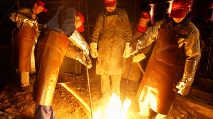 Glockengiesser giessen eine rund 1,5 Tonnen schweren Glocke, deren Form sich im Boden befindet. Aus dem Eingussloch schlagen Flammen.