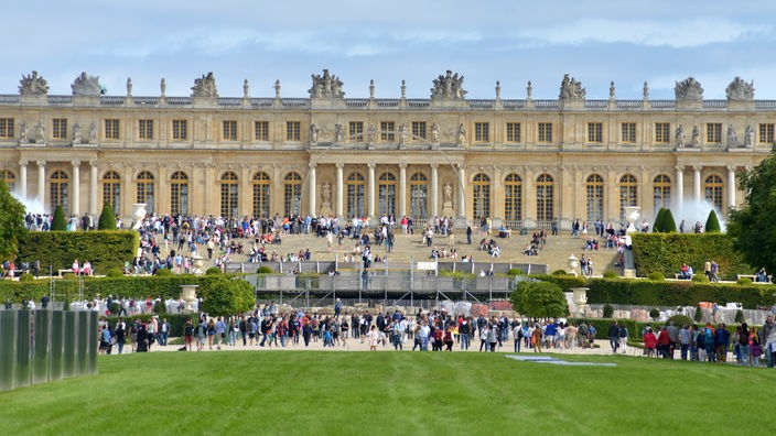 Zahlreiche Touristen tummeln sich auf dem symmetrischen Vorplatz vor dem Schloss, dass sich über die ganze Bildbreite erstreckt 