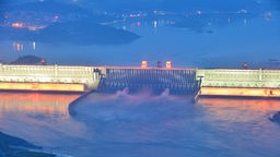 Luftaufnahme des Drei-Schluchten-Staudamms in China. Ein rieisger beleuchteter Staudamm zieght sich durch ein Gewässer, das große Wassermengen führt.