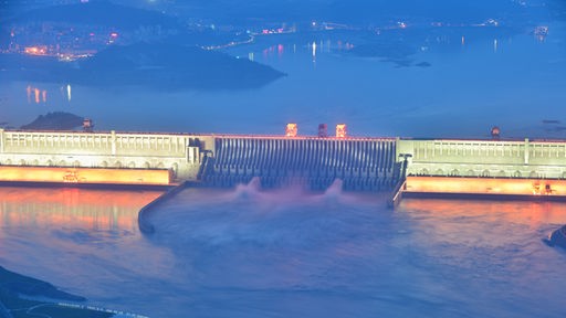 Luftaufnahme des Drei-Schluchten-Staudamms in China. Ein rieisger beleuchteter Staudamm zieght sich durch ein Gewässer, das große Wassermengen führt.
