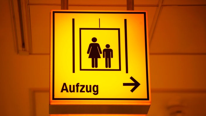 Ein gelbes Schild mit einer stilisierten Frau und einem Kind weist den Weg zu einem Fahrstuhl