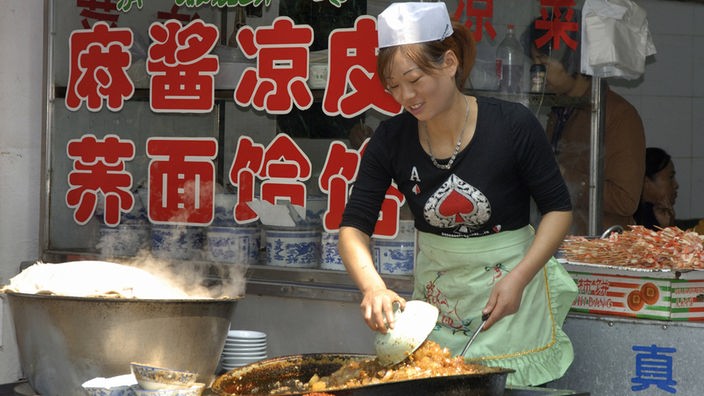 Eine junge Frau bereitet ein Gericht in einer riesigen Pfanne zu. Sie steht vpr einem Laden, das Schaufenster ist mit großen chinesischen Schriftzeichen beklebt. 
