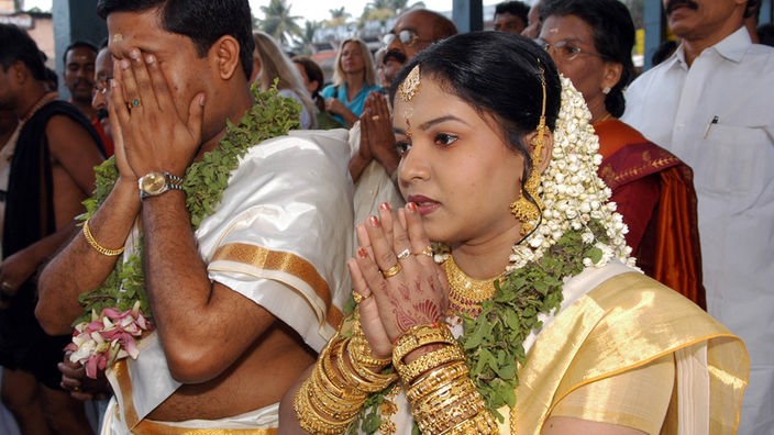 Braut und Bräutigam tragen traditionelle indische Gewänder, sie stehen nebeneinander und beten, hinter ihnen ist die Hochzeitsgesellschaft versammelt. 