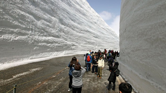 Einige Menschen laufen über eine Straße, zwischen 17 Meter hohen Schneewänden zu beiden Seiten