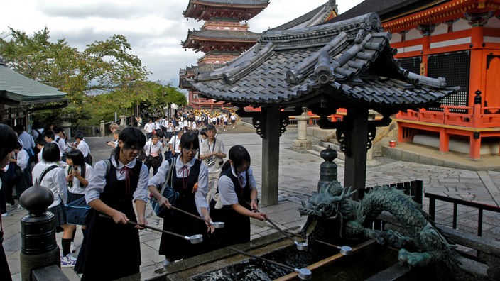 Drei japanischische Schülerinnen in Uniform reinigen sich die Hände an einem Wasserbecken vor einem Tempel.