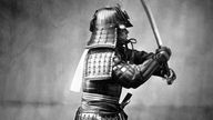 Das schwarz-weiß-Foto zeigt einen Samurai in voller Rüstung, der sein Schwert schwingt