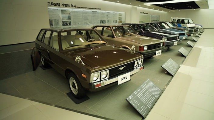 Im Museum stehen mehrere Modelle des Hyundai Pony in einer Reihe, davor sind Tafeln mit den dazugehörigen Daten zu erkennen. 