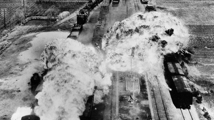 Luftbild von Zügen auf zwei Bahngleisen, die nach einem Bombenangriff in Flammen aufgehen.