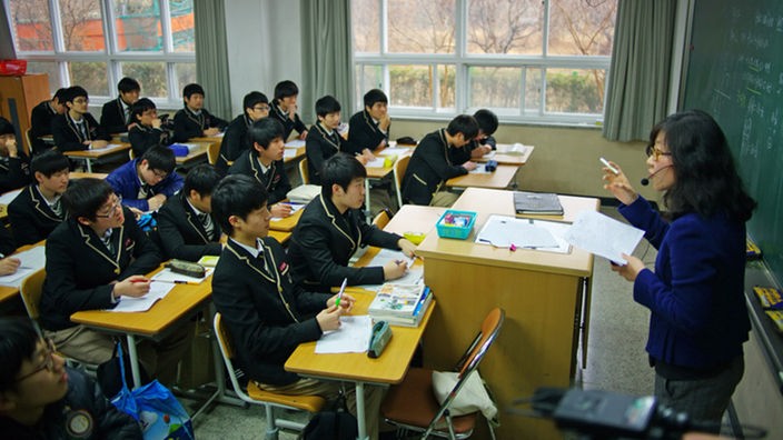 In einer koreanischen Schulklasse sitzen Jugendliche mit Schuluniform, vor ihnen steht eine Lehrerin mit einem Headset.