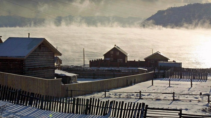 Einige Holzhäuser im Winter am Baikalsee im Gegenlicht. Die Oberfläche des Sees dampft.
