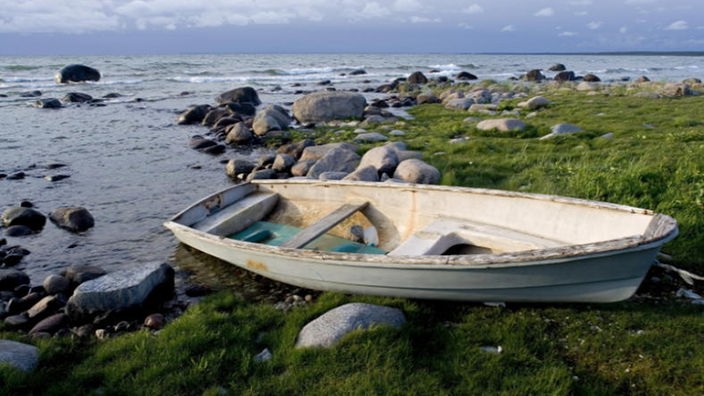 Ein hölzernes Ruderboot liegt im Gras der Küste, aus dem Meer ragen zahlreiche Felsen.