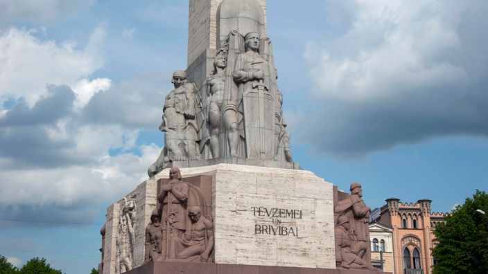 Das Foto zeigt das Freiheitsdenkmal in Riga. Am Fuß des Monolithen aus Stein sind verschiedene Figuren abgebildet