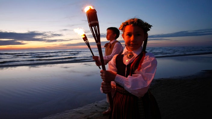 Traditionelles Mittsommernachtsfest am Strand von Jurmala: Zwei Kinder mit Fackeln an einem Strand vor der untergehenden Sonne.