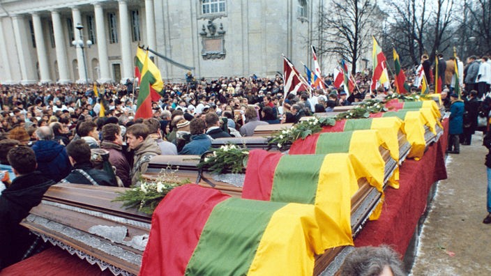 Särge, aufgereiht und in litauische Flaggen gehüllt, darum herum viele Menschen.