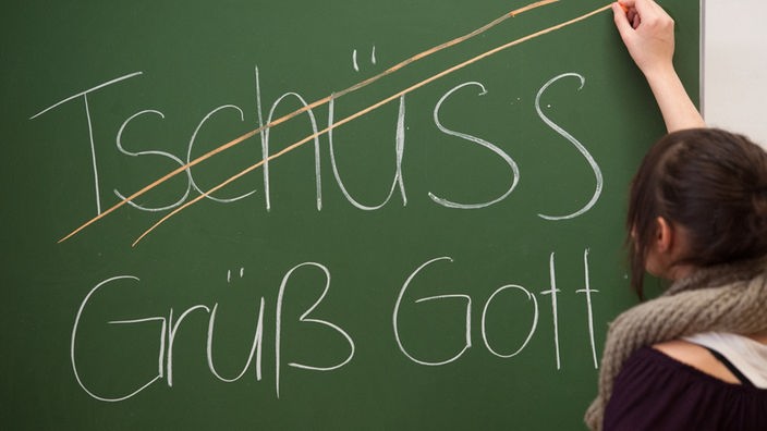 Eine Schülerin streicht das Wort Tschüss auf einer Tafel durch, darunter steht stattdessen Grüß Gott