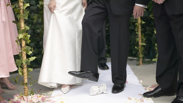 Nach jüdischer Tradition zertritt der Bräutigam mit dem Schuh ein Glas. Man sieht den Fuß des Bräutigams sowie die Füße der umstehenden Gäste und der Braut.