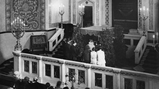 Das Schwarzweiß-Foto zeigt eine Trauung in einer Synagoge. Das Brautpaar steht unter einem Baldachin. Im Gestühl sitzen Hochzeitsgäste und schauen zu.