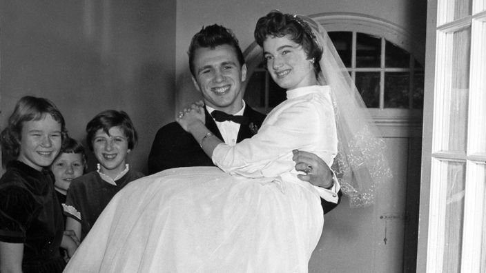 Ein Schwarzweiß-Foto zeigt einen Bräutigam, der die Braut über die Türschwelle einer Wohnung trägt. Links sieht man drei lachende Kinder.