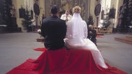 Ein Brautpaar während der kirchlichen Trauung von hinten. Wenn man zwischen den Brautleuten hindurch schaut, kann man den Pfarrer sehen.