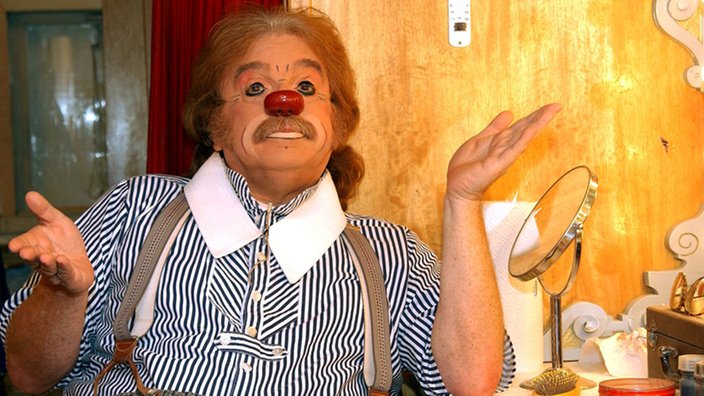 Bernhard Paul schminkt sich vor einem Spiegel als Clown Zippo. Er trägt ein blauweißes Hemd mit weißem Kragen und eine grau karierte Hose.