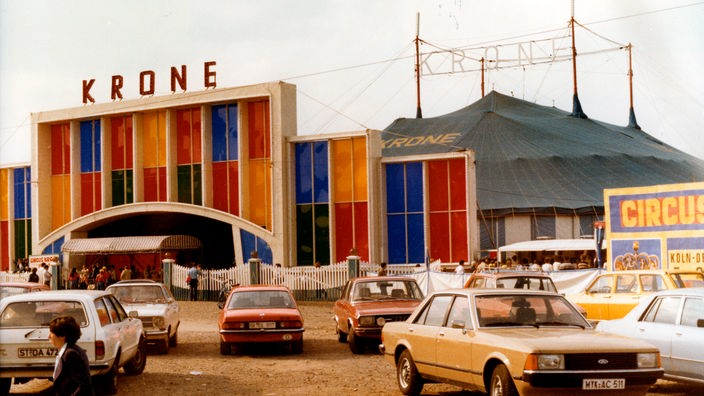 Blick auf ein Zirkuszelt mit großem Vorbau, auf dem 'Circus Krone' steht. Davor stehen einige Autos auf einem sandigen Parkplatz.