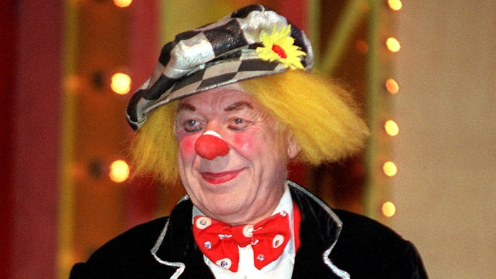 Clown Oleg Popow steht mit roter Nase, karierter Mütze, großer Fliege und Jacket in der Manege.