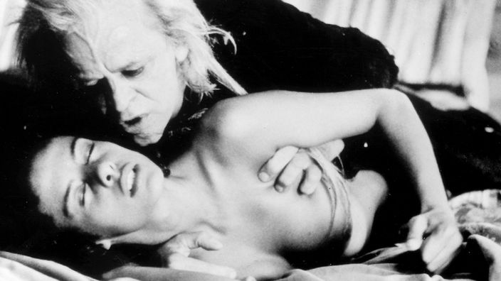 Schwarzweiß-Filmszene: Klaus Kinski als Vampir beugt sich über ein im Bett liegendes hübsches weibliches Opfer.