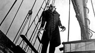 Schwarzweiße Filmszene: Dracula mit Glatze, schwarzem Mantel, großen Ohren und sehr langen Fingernägeln auf einem Schiff