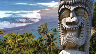 Eine antike polynesische Holzstatue vor einem Palmenstrand auf Hawaii.