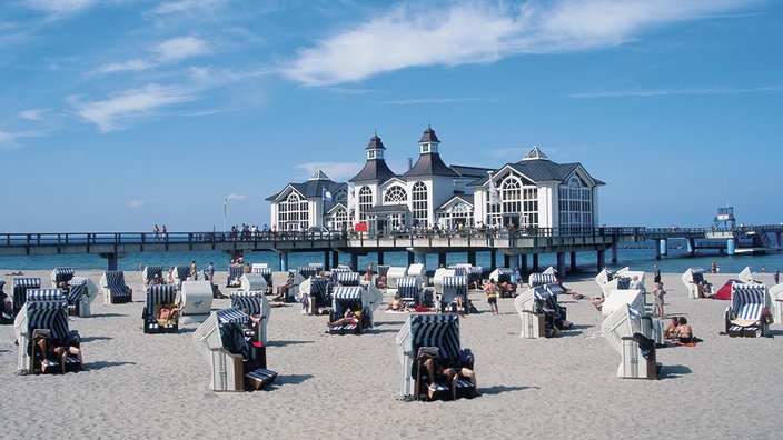 An einem Sandstrand sitzen Menschen in Strandkörben. Im Hintergrund steht ein weißes Haus mit roten Türmen auf Stelzen im Wasser. | Bildquelle: dpa An einem Sandstrand sitzen Menschen in Strandkörben. Im Hintergrund steht ein weißes Haus mit roten Türmen auf Stelzen im Wasser.