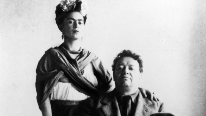Schwarz-weiß-Fotografie von Frida Kahlo und ihrem Mann Diego Rivera; er sitzt auf einem Stuhl, seinen Hut in der Hand haltend. Sie steht hinter ihm und legt ihm eine Hand auf die Schulter. | Bildquelle: picture alliance / Everett Collection Schwarz-weiß-Fotografie von Frida Kahlo und ihrem Mann Diego Rivera; er sitzt auf einem Stuhl, seinen Hut in der Hand haltend. Sie steht hinter ihm und legt ihm eine Hand auf die Schulter.