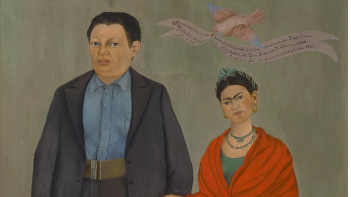 Fotografie des Werks „Frida und Diego Rivera“ von 1931. Auf dem Gemälde sind Frida Kahlo und Diego Rivera am Tag ihrer Hochzeit zu sehen; er trägt einen grauen Anzug, sie ein grünes Kleid und einen roten Schal.