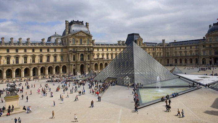 Blick in den Innenhof des Louvre in Paris. Zu sehen sind die Fassade des Museums, eine große Glaspyramide und etliche Besucher